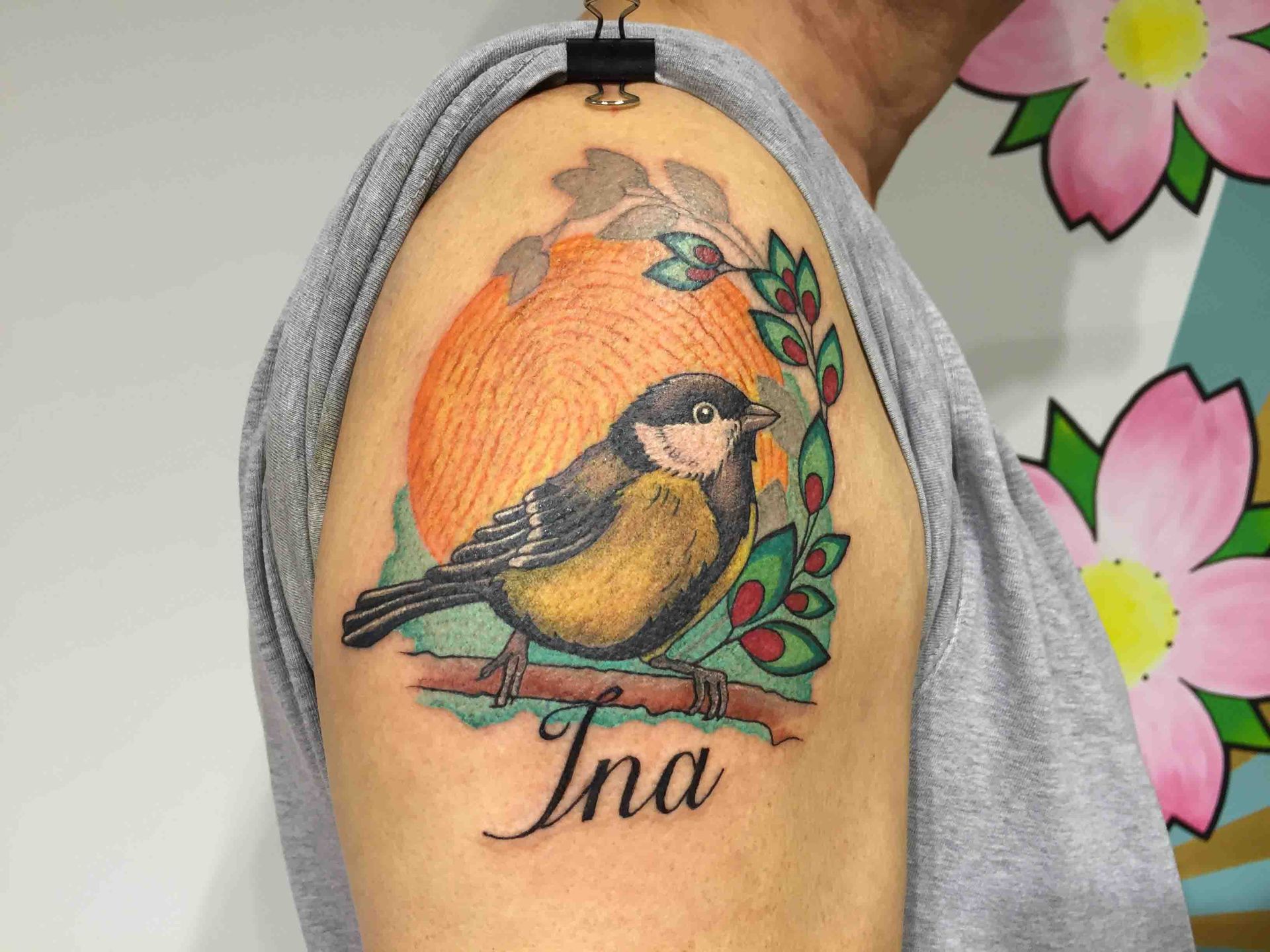 Koolmees op een tak voor een oranje zon, met de naam Ina eronder, op een schouder