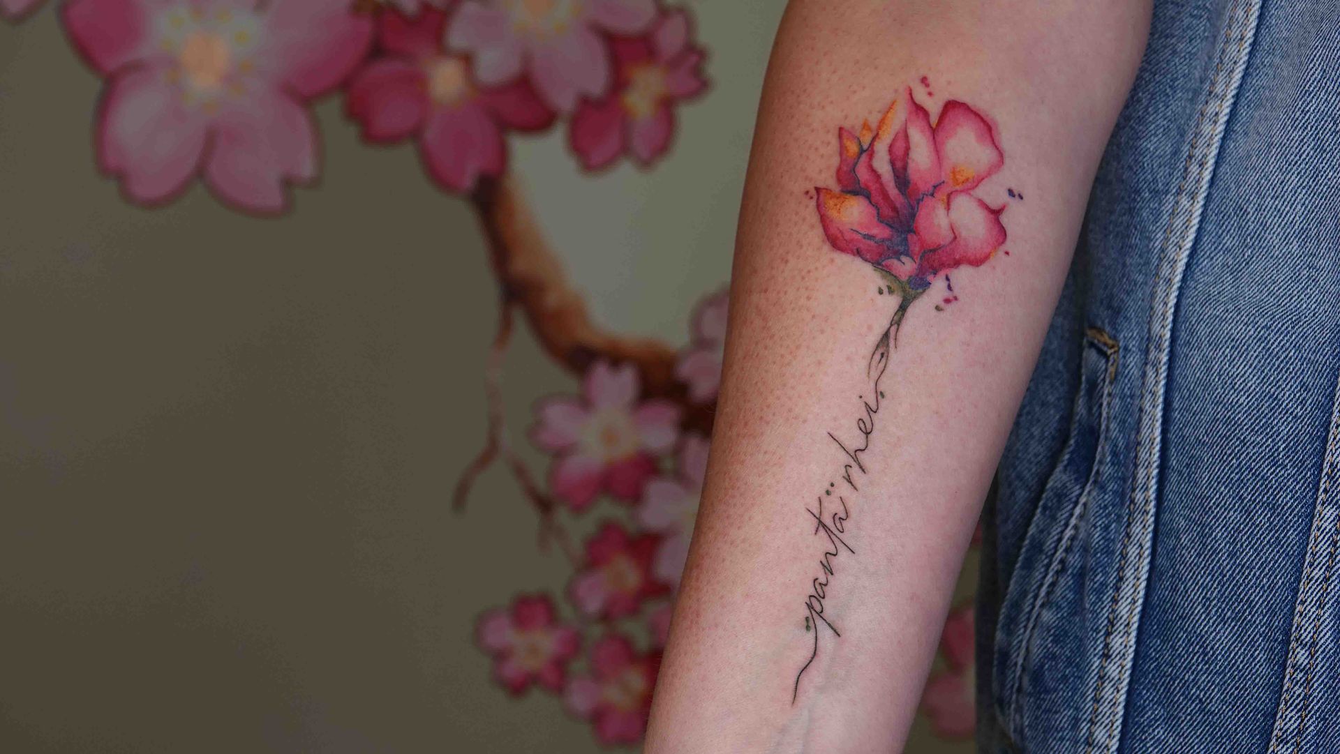 Roze bloem in waterverfstijl op een onderarm, met een dunne handgeschreven tekst eronder