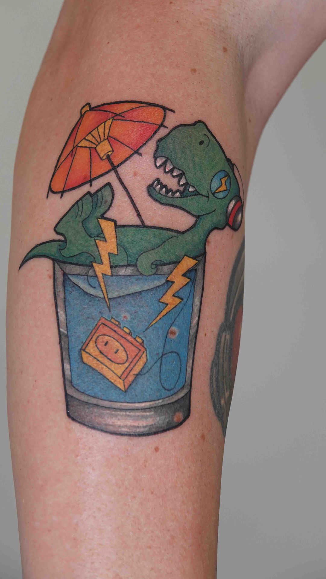 Tekenfilmdinosaurus in een drinkglas met parasol en bliksemschichten, op een bovenarm